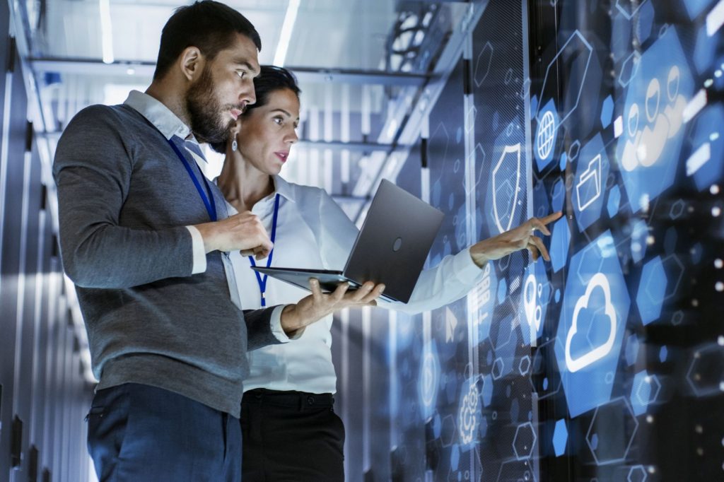 Uma mulher e um homem com um notebook nas mãos em uma sala de servidores de computador observam uma parede com ícones representando elementos tecnológicos