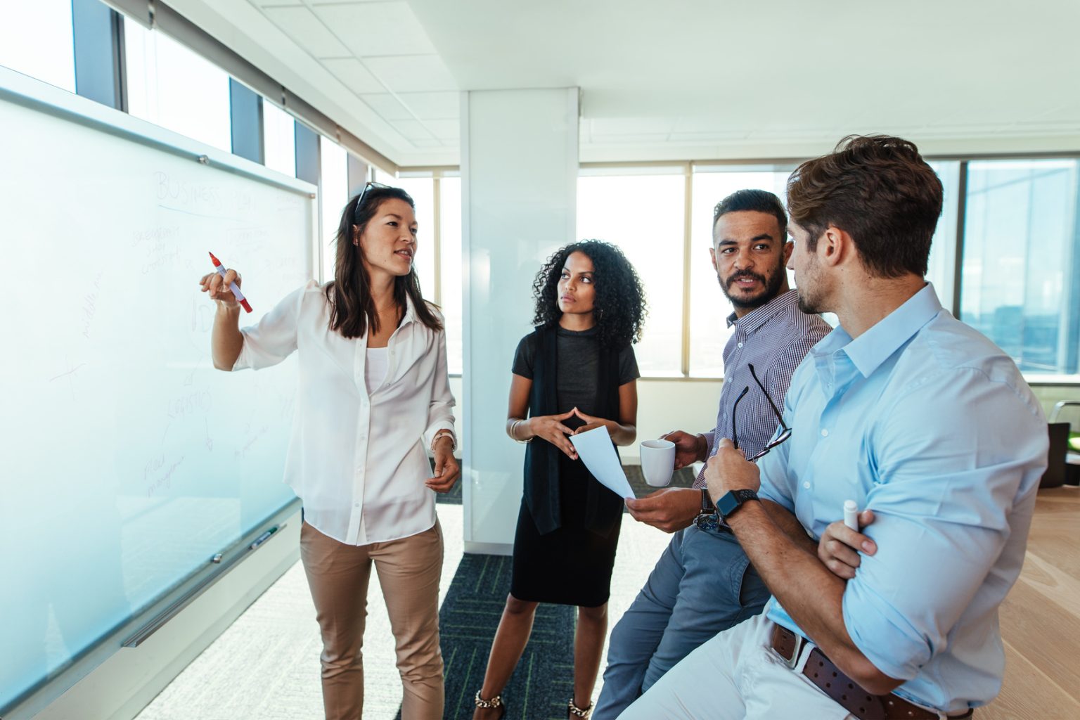 Mulher em frente a uma lousa faz anotações e debate ideias, enquanto colegas de trabalho observam