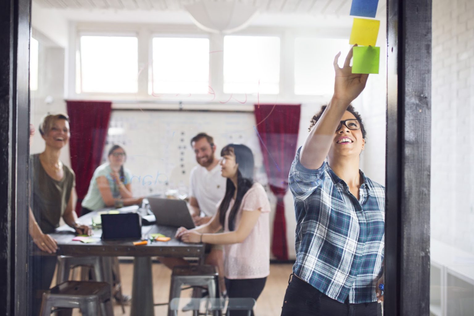 Grupo de trabalhadores em escritório debatendo ideias. À frente, uma mulher negra cola post-its numa lousa