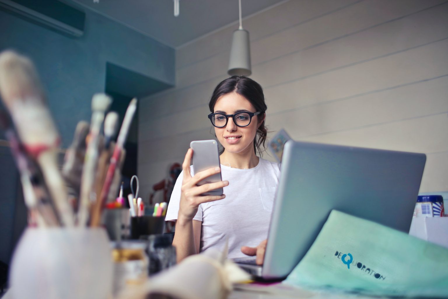 Jovem mulher observa o celular sentada em sua mesa de trabalho com diversas canetas e um notebook