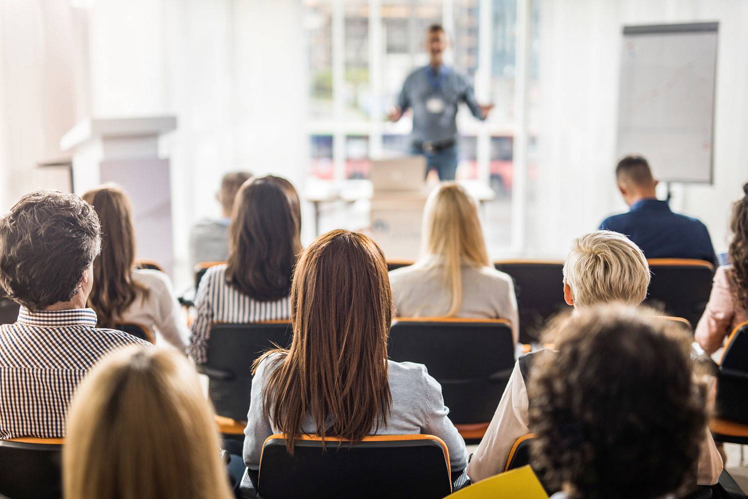 Sala de aula cheia, com pessoas de diversas etnias e idades, observam um professor em frente a um púlpito